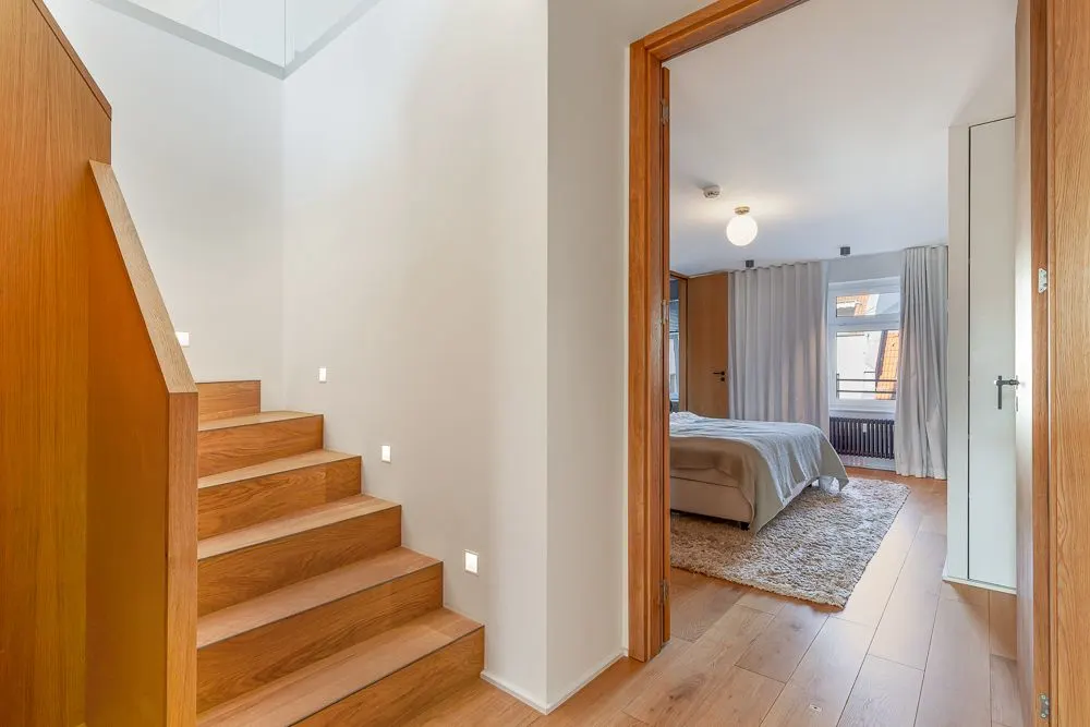 Flur mit Holztreppe und Blick in ein Schlafzimmer mit Bett und großem Fenster.