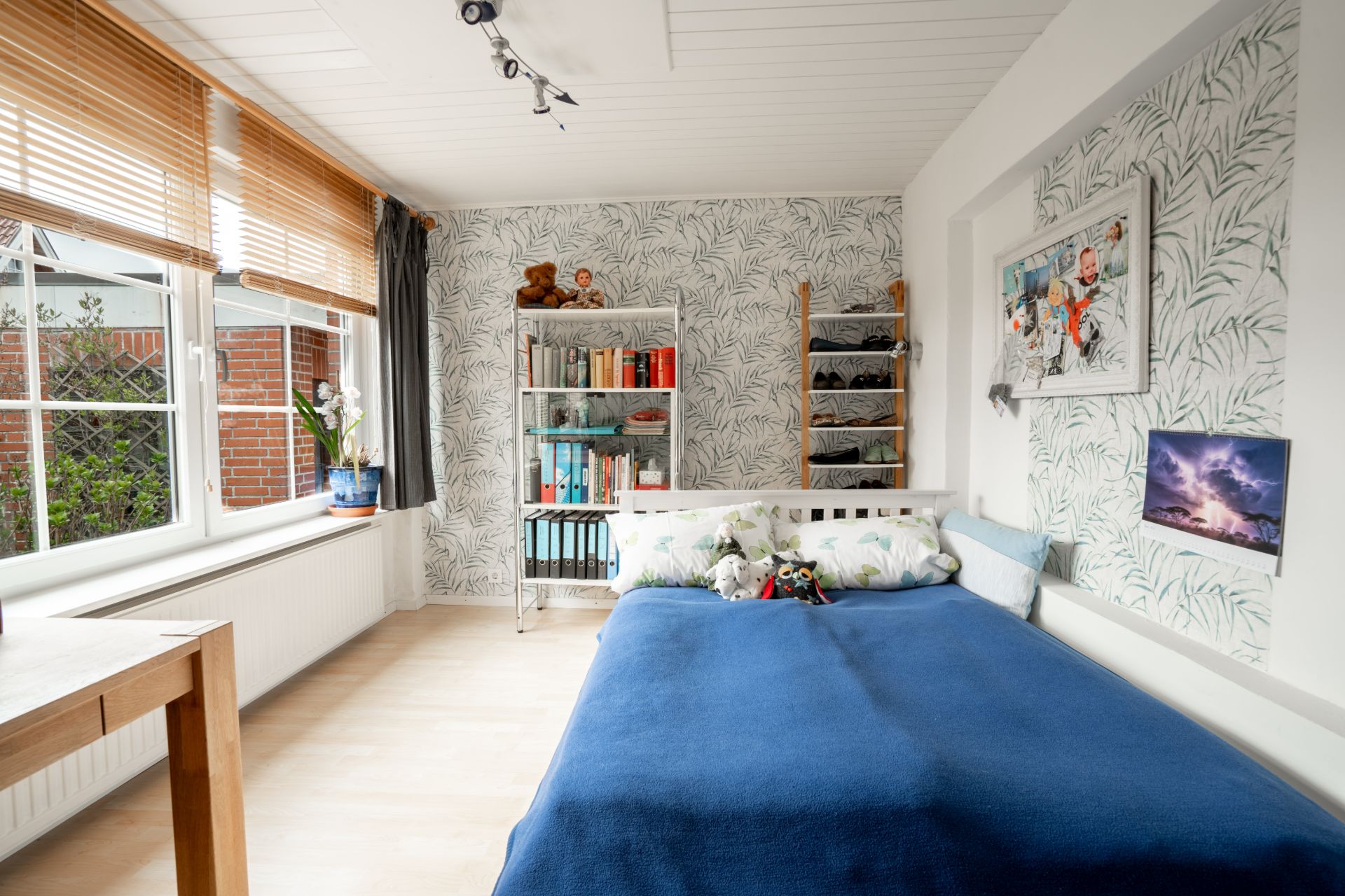 Helles Schlafzimmer mit großem Fenster, Bett mit blauer Decke und Regal mit Büchern und Kuscheltieren.