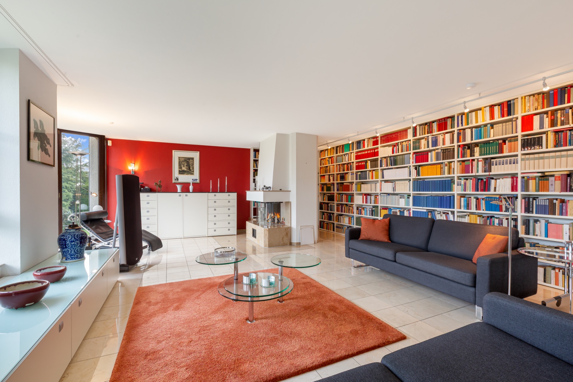 Helles Wohnzimmer mit Bücherwand, zwei Sofas, Glastischen und rotem Teppich.