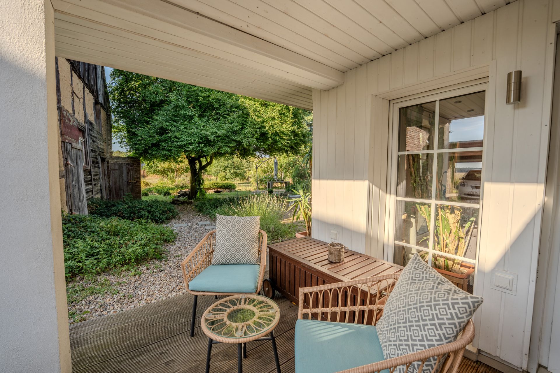 Überdachte Terrasse mit zwei Stühlen, kleinem Tisch und Blick in einen Garten mit Bäumen.