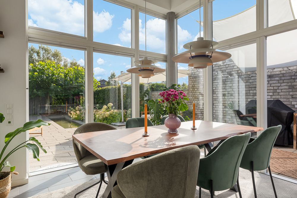 Essbereich mit Holztisch, grünen Stühlen und großen Fenstern mit Blick auf Garten.