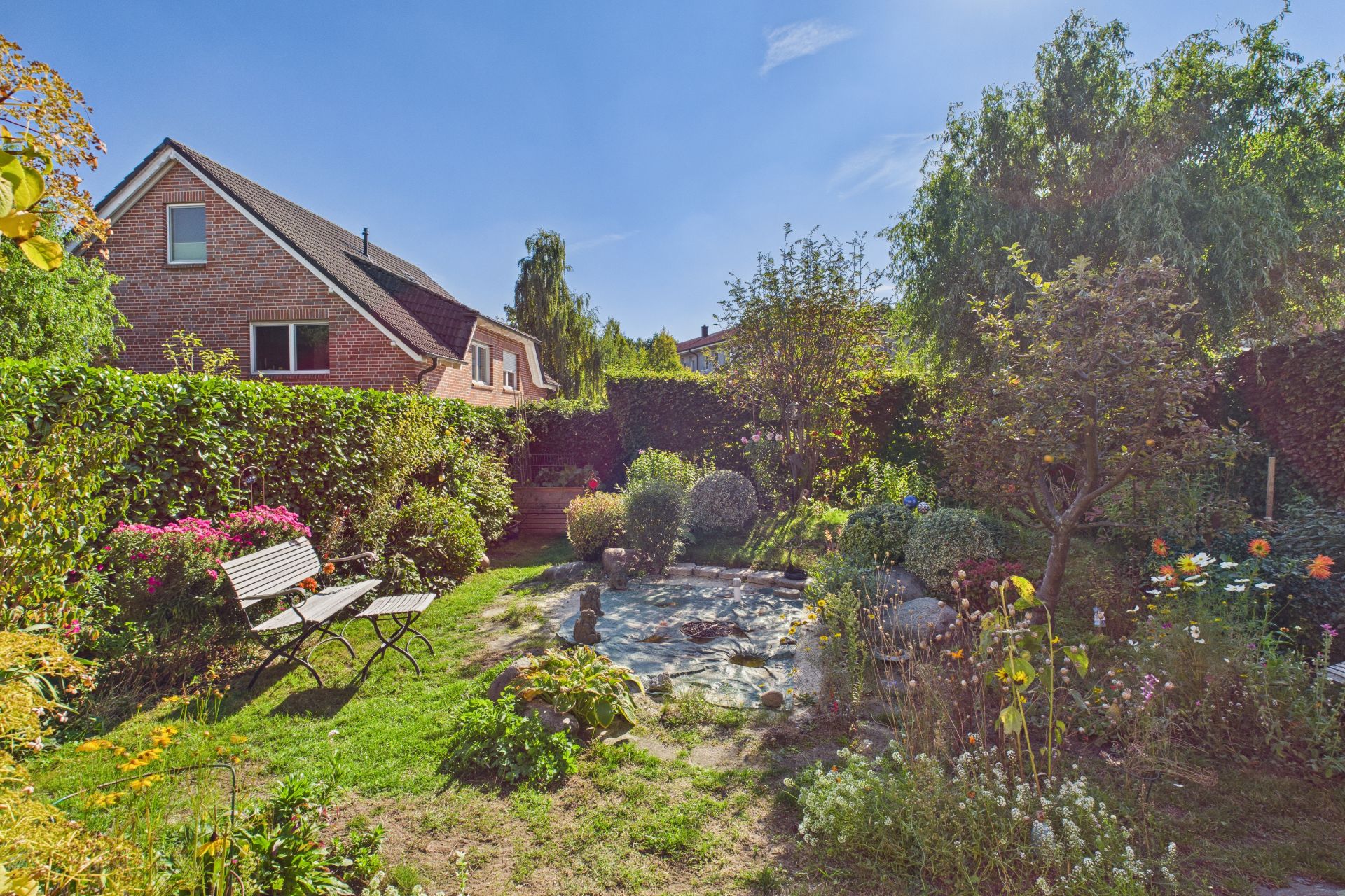 Grüner Garten mit Teich, Blumen, Sträuchern und einer Bank, Haus im Hintergrund.