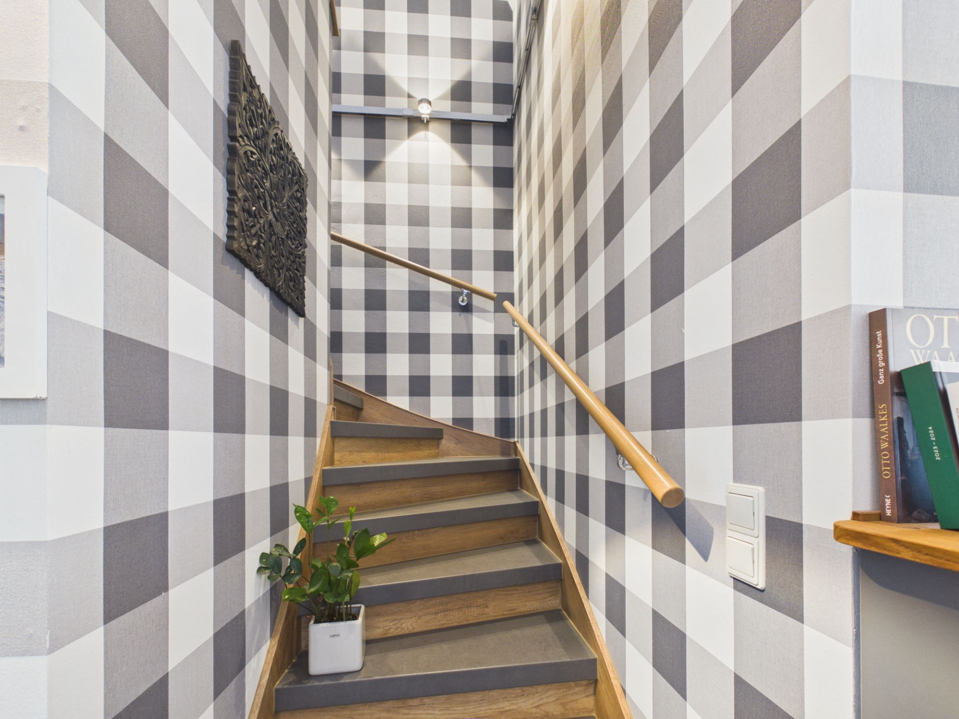 Treppe mit Holzstufen und karierten Wänden, Pflanze im Topf, Handlauf und Wandbeleuchtung.