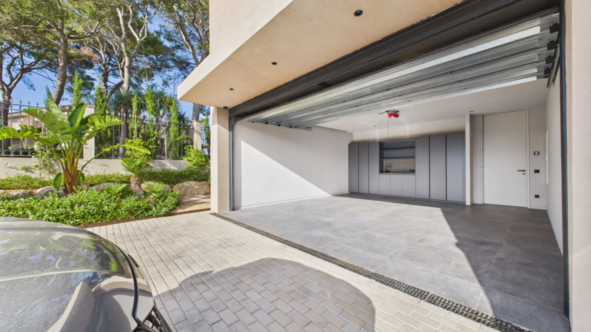 Geöffnete Garage mit grauem Boden und Schränken an der Rückwand, daneben Pflanzen und gepflasterter Zufahrt.
