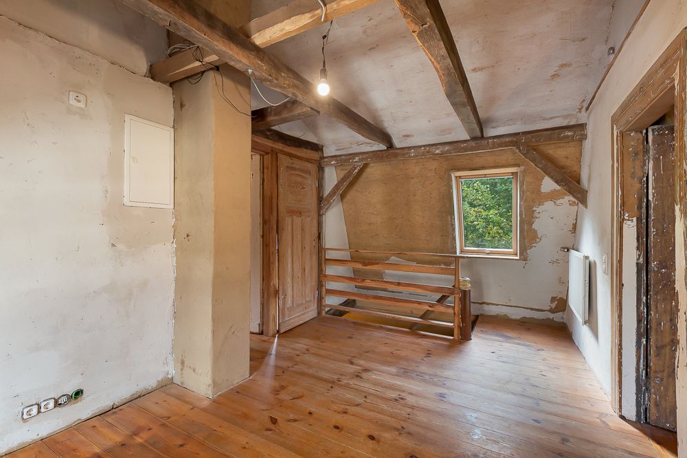 Ein Zimmer mit Holzboden, sichtbaren Holzbalken, Fenster und Geländer vor einer kleinen Treppe.
