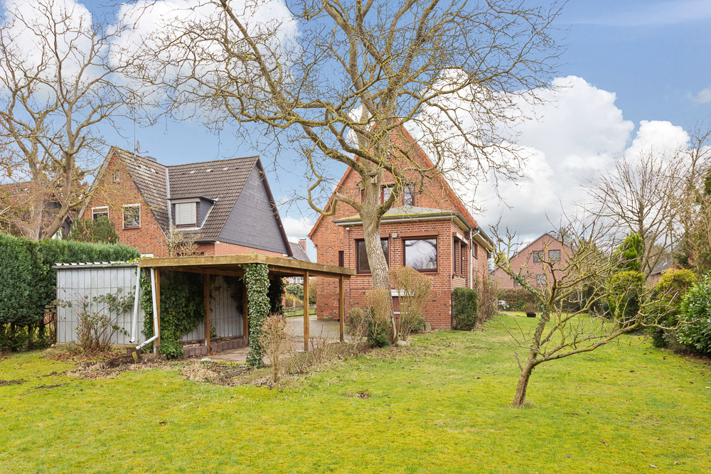 Garten mit Rasenfläche, Baum, überdachtem Unterstand und rotem Backsteinhaus im Hintergrund.