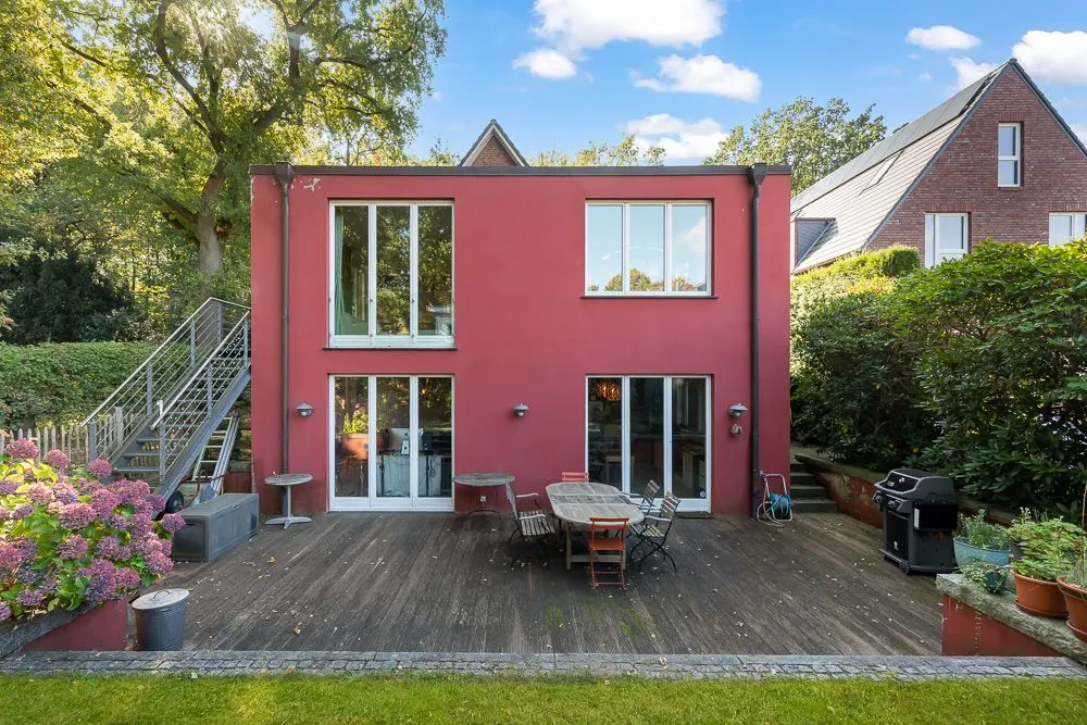 Rotes Wohnhaus mit großer Holzterrasse, Gartenmöbeln und Grill im grünen Garten.