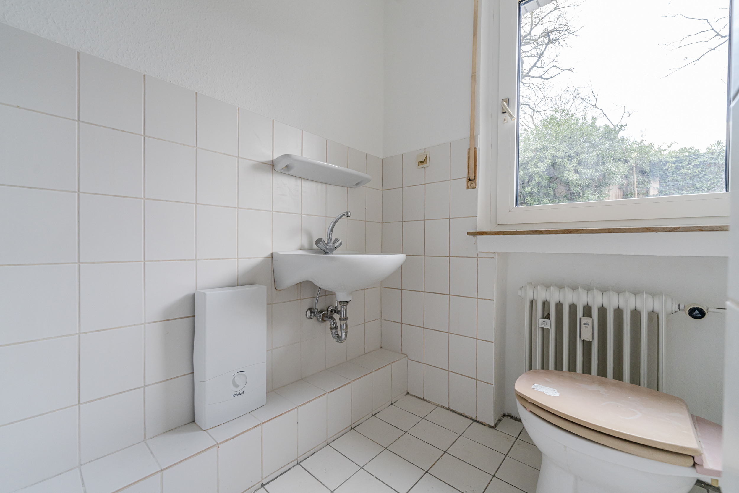 Kleines Badezimmer mit Waschbecken, Toilette, Heizung und Fenster, weiße Fliesen an Wand und Boden.