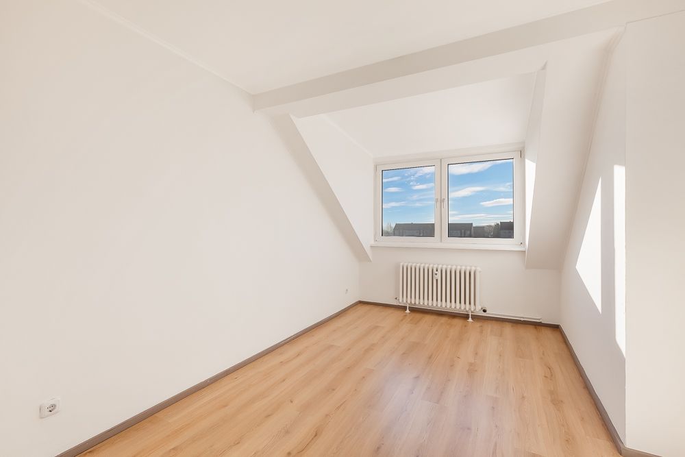 Helles Zimmer mit Holzfußboden, weiß gestrichenen Wänden und großem Fenster unter einer Schräge.