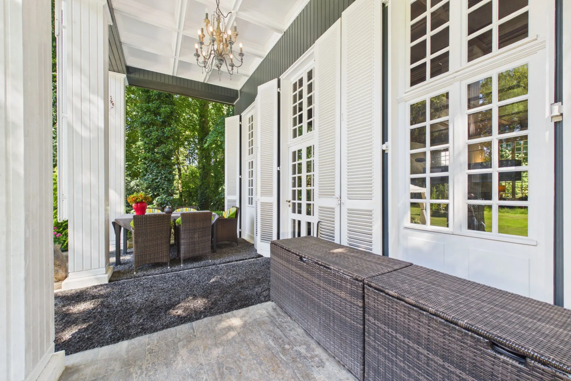 Überdachte Terrasse mit weißen Türen, Rattanmöbeln und Esstisch, Blick auf grünen Garten.