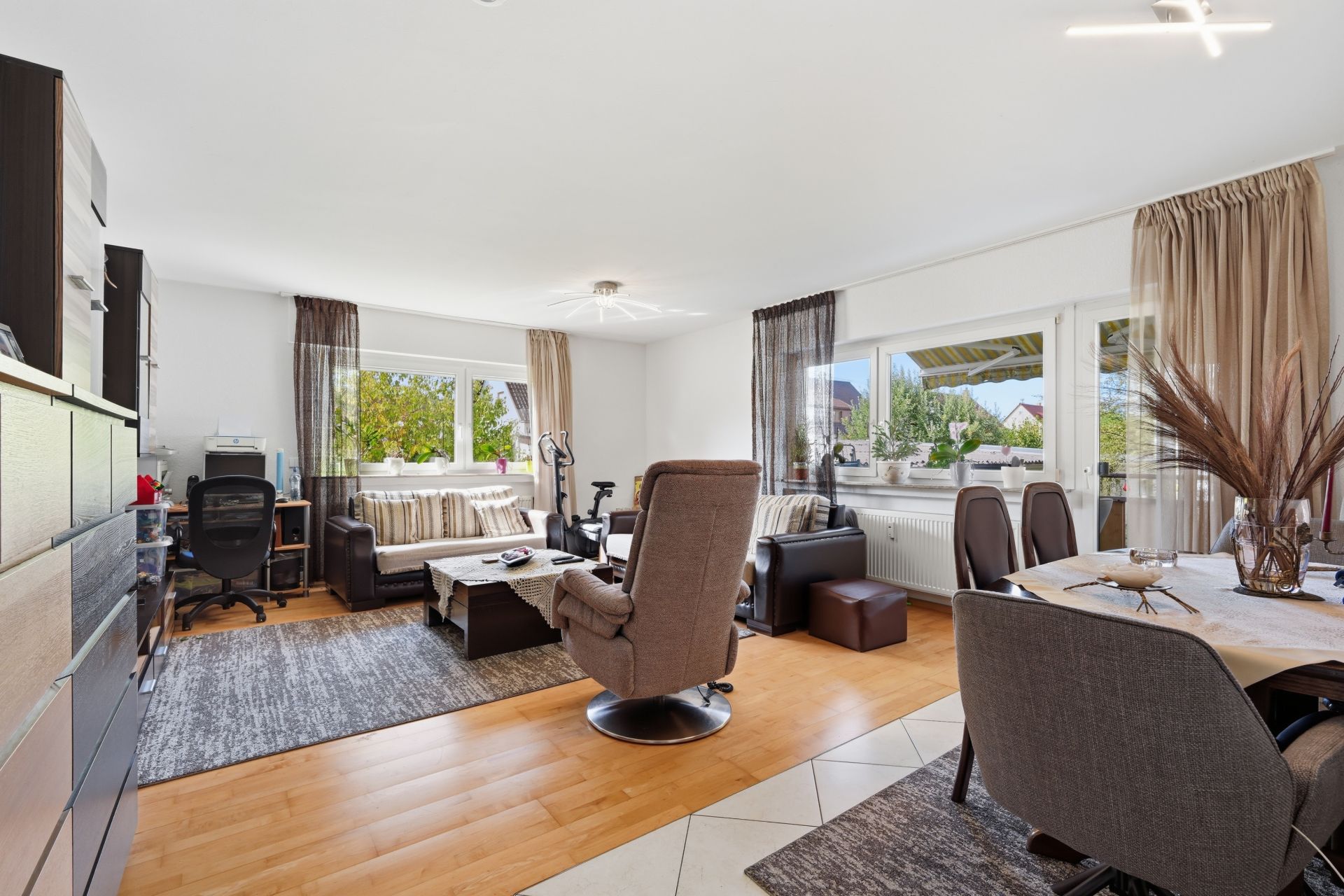 Helles Wohnzimmer mit Sofa, Sessel, Esstisch und Schreibtisch, große Fenster mit Vorhängen.
