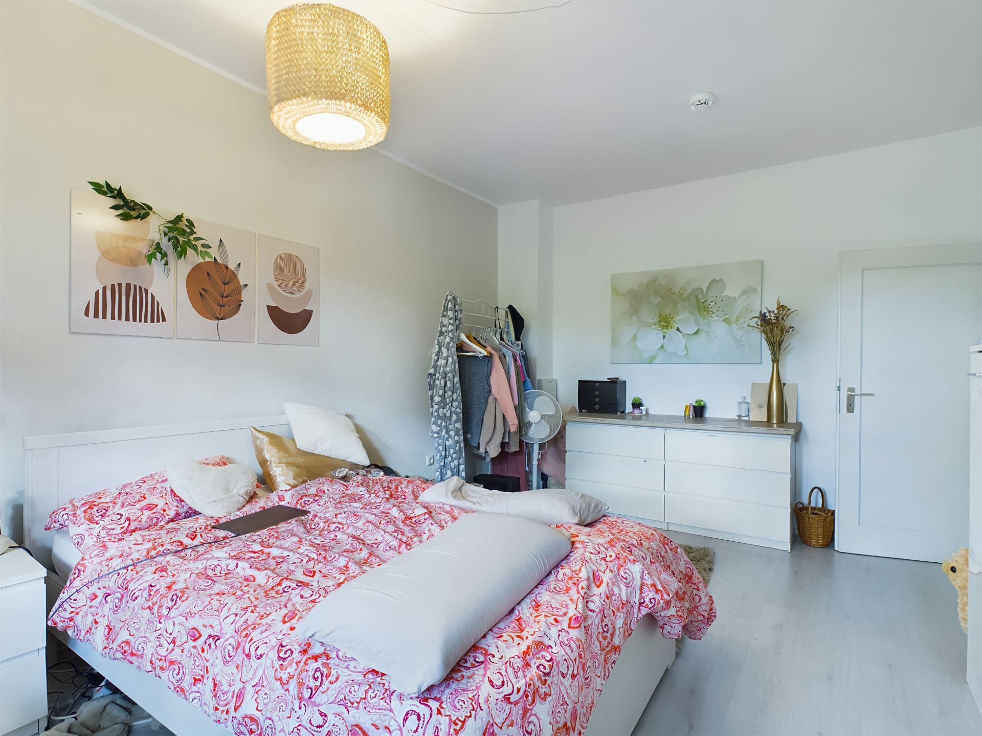 Helles Schlafzimmer mit Doppelbett, bunter Bettwäsche, Kommode und dekorativen Bildern an weißen Wänden.