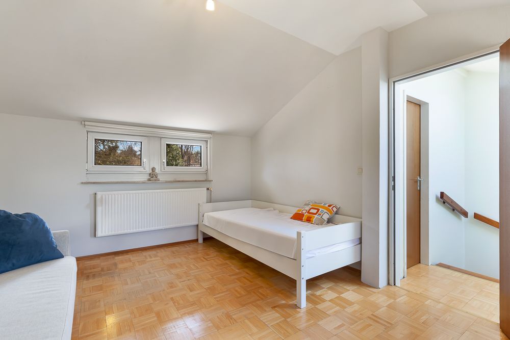 Helles Schlafzimmer mit Einzelbett, Parkettboden und zwei Fenstern, Tür zum Flur offen.