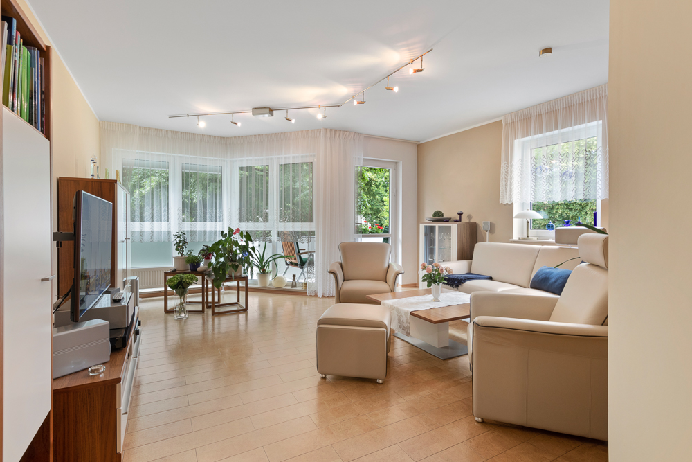 Helles Wohnzimmer mit Ledersofas, Fernseher, Pflanzen und großen Fenstern mit Vorhängen.