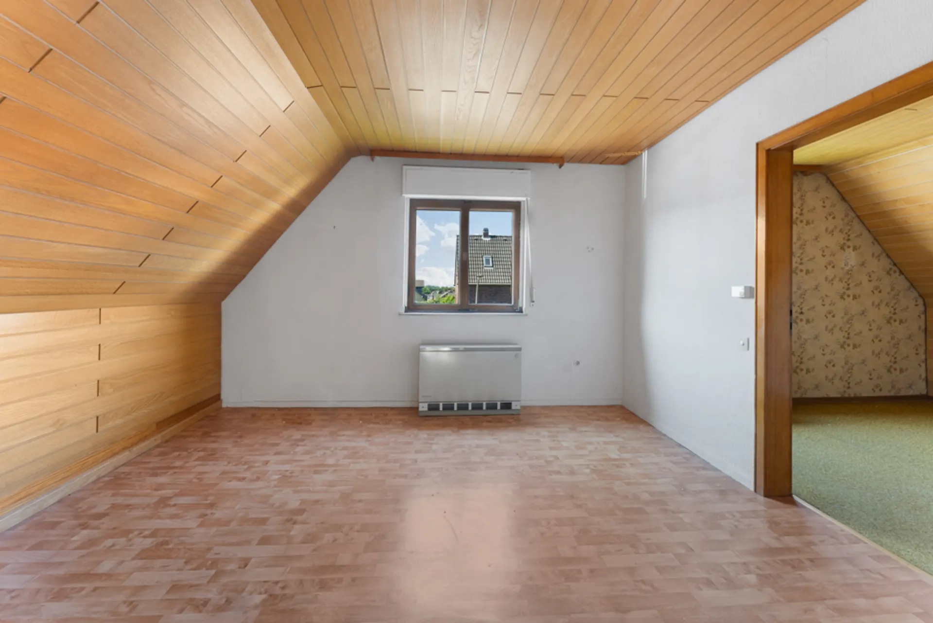 Leerer Raum mit Holzdecke, schräger Wand, Fenster und offenem Durchgang zu weiterem Zimmer.