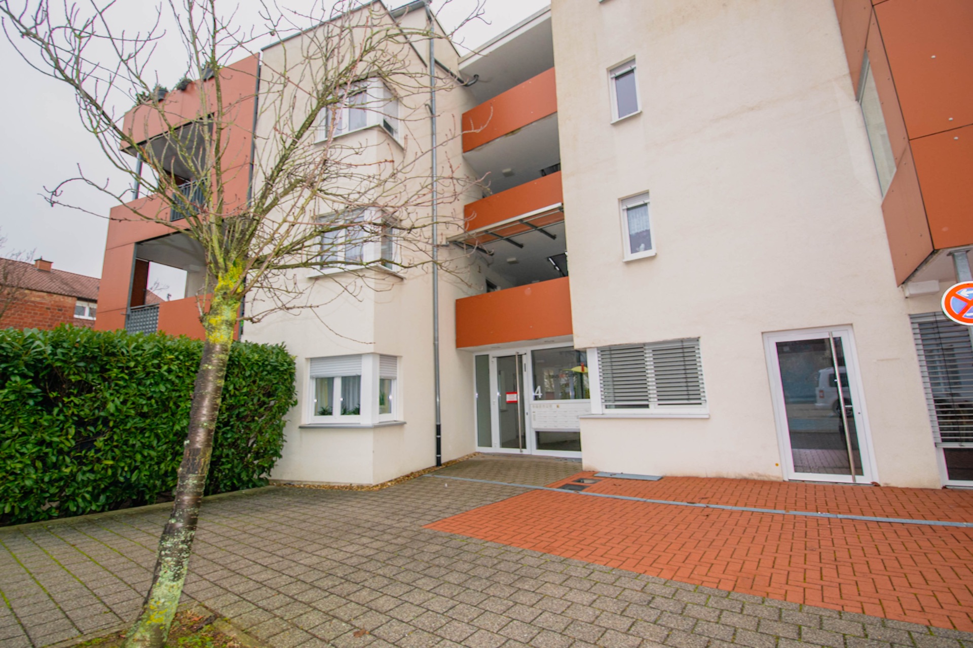 Modernes Wohnhaus mit weißer Fassade, orangenen Balkonen und gepflastertem Eingangsbereich.