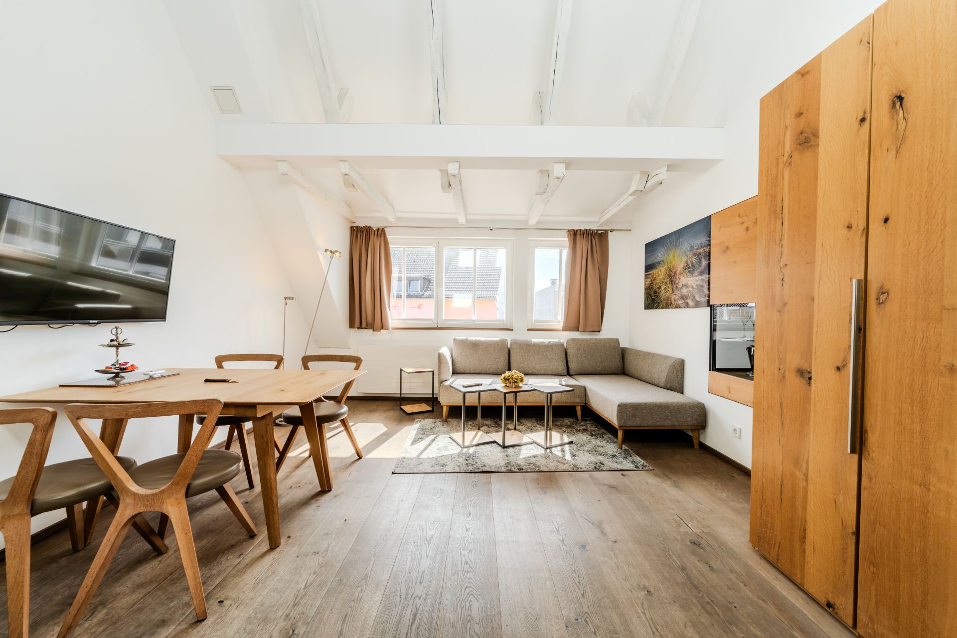 Helles Wohnzimmer mit Ecksofa, Esstisch, Fernseher und Holzschrank auf Holzboden.