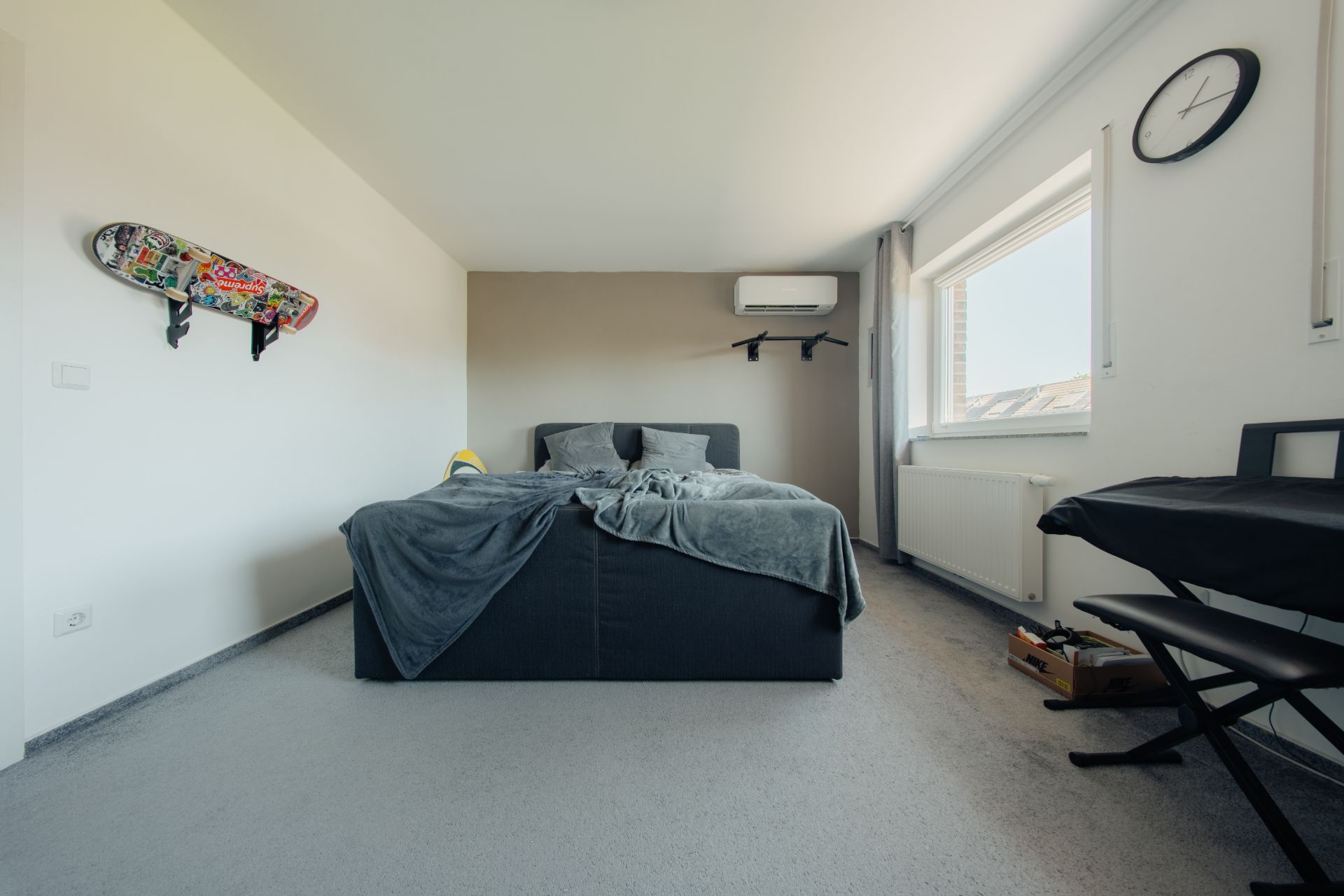 Helles Schlafzimmer mit grauem Bett, Skateboard an der Wand und Fenster mit Vorhang.