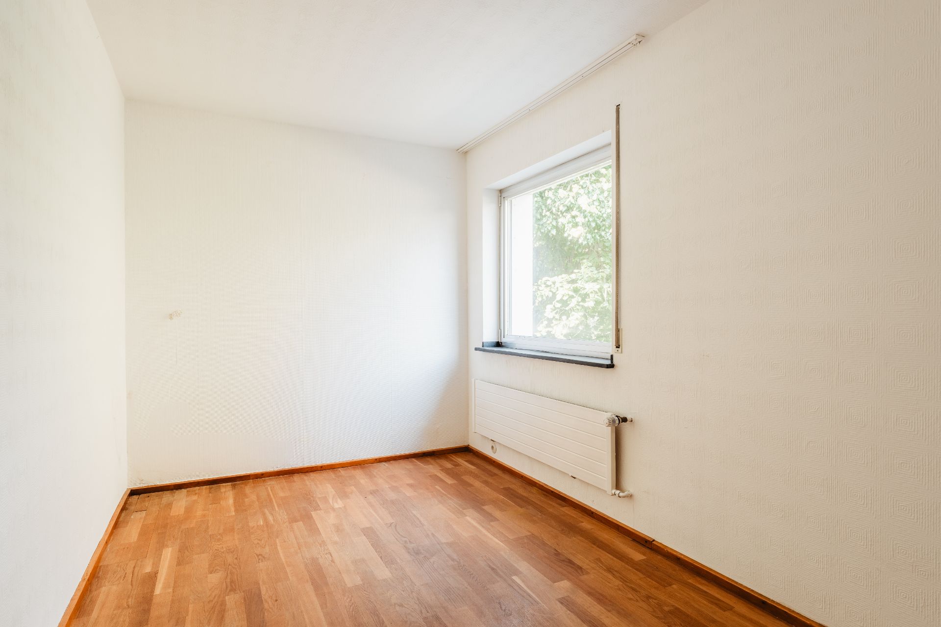 Leerer Raum mit Holzboden, weißer Wand und Fenster mit Blick ins Grüne.