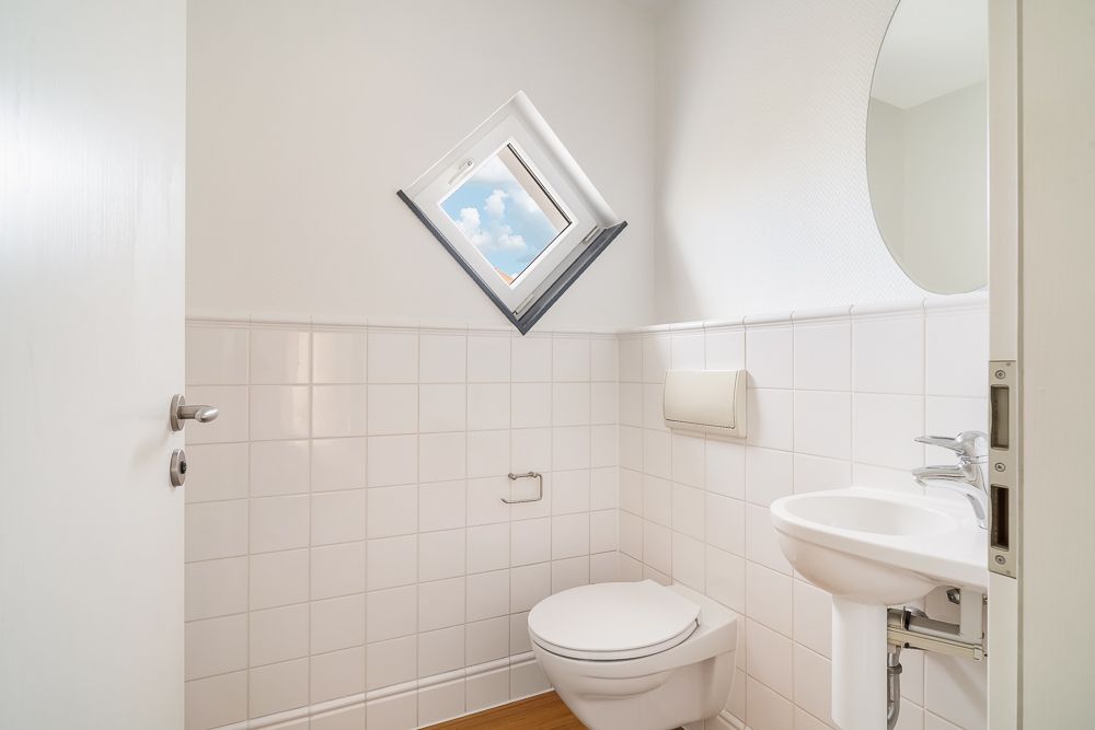 Helles Badezimmer mit Toilette, Waschbecken, rundem Spiegel und kleinem Fenster.