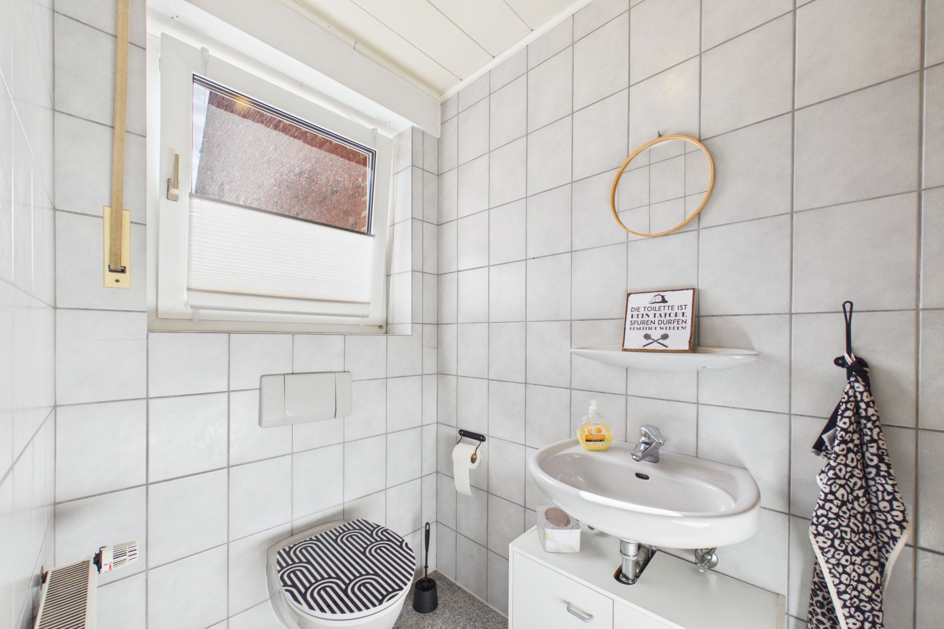 Helles Badezimmer mit Fenster, Toilette, Waschbecken, Handtuch und Spiegel an der gefliesten Wand.