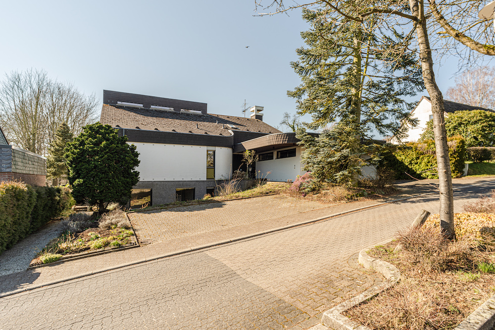 Modernes Wohnhaus mit schrägem Dach, Einfahrt und umliegenden Bäumen bei sonnigem Wetter.
