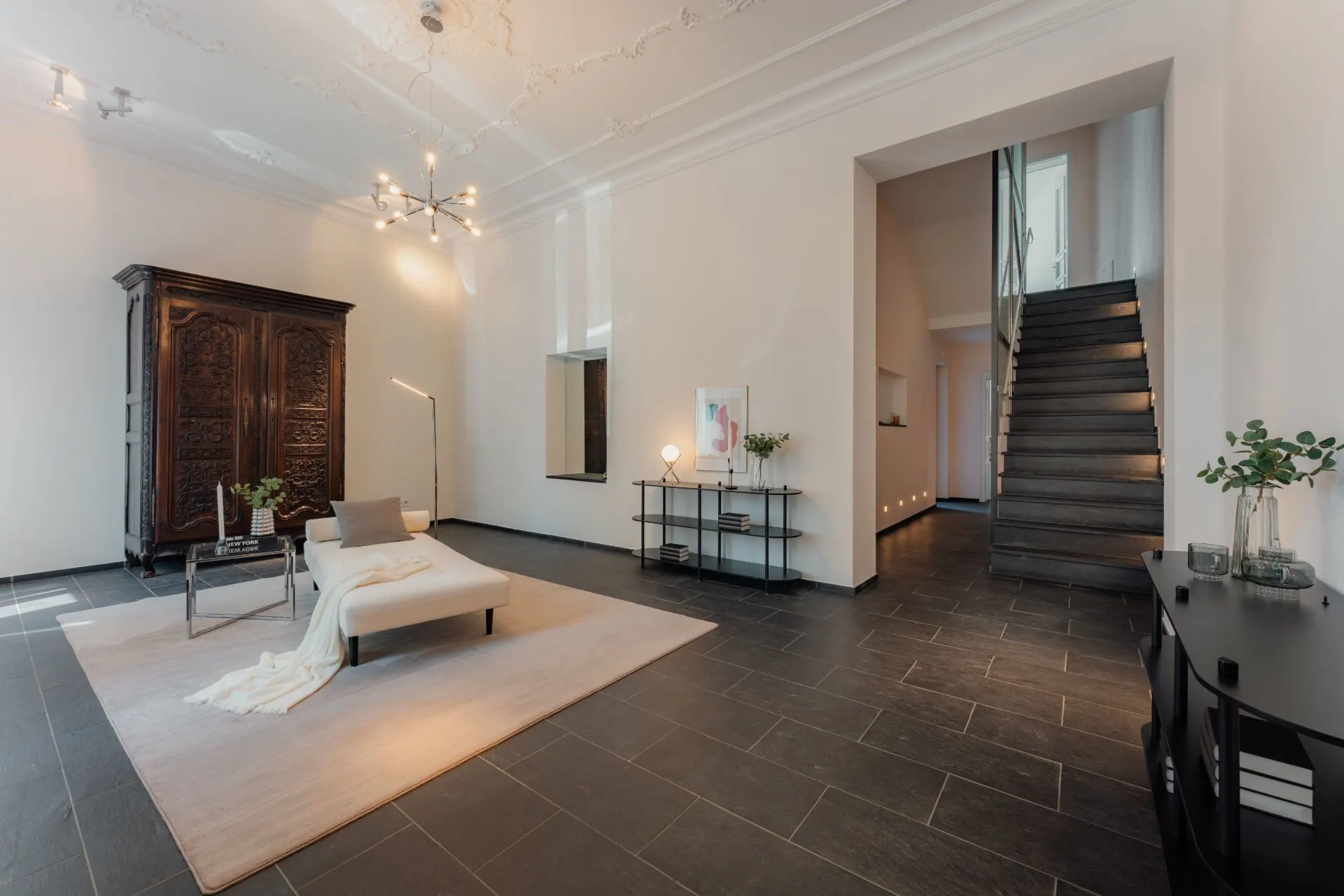 Modernes Wohnzimmer mit Sofa, Teppich, dunklem Schrank und Treppe, dekoriert mit Pflanzen und Kunst.