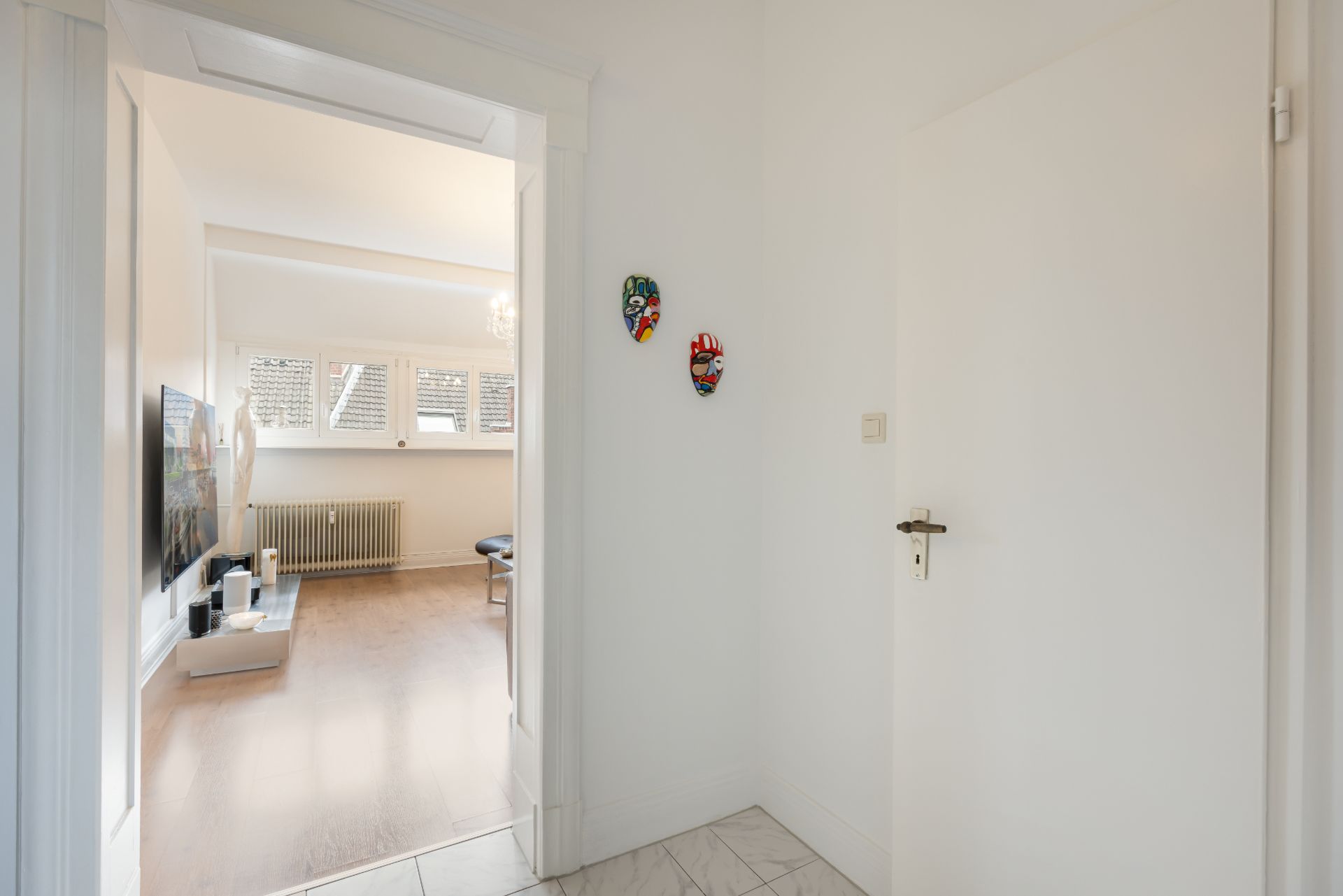 Blick vom Flur in ein Wohnzimmer mit Fernseher, Holzfußboden und zwei Masken an der Wand.