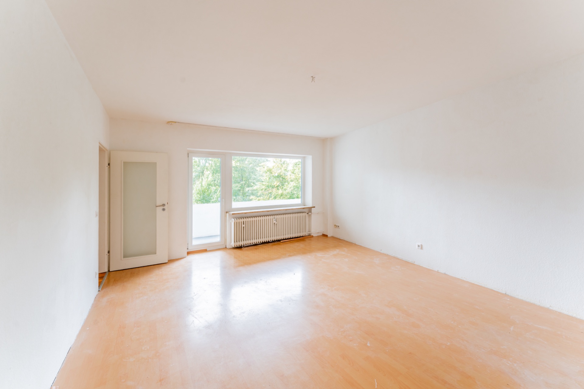 Leerer Raum mit hellem Boden, weißer Wand, Tür und großem Fenster mit Heizung darunter.