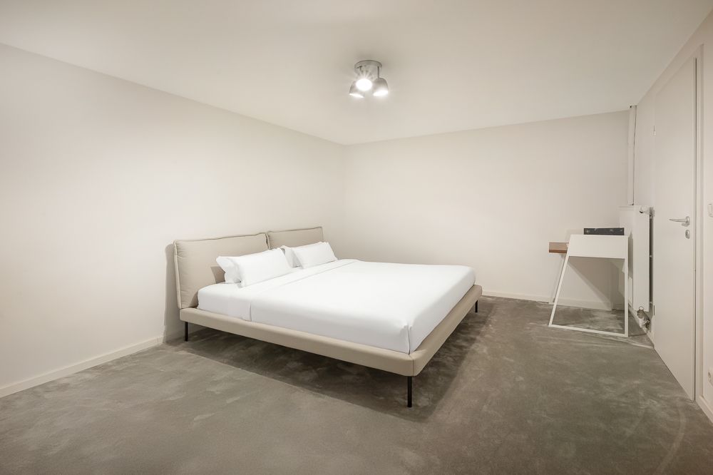 Schlafzimmer mit Doppelbett, Nachttisch und Schreibtisch auf grauem Teppichboden.