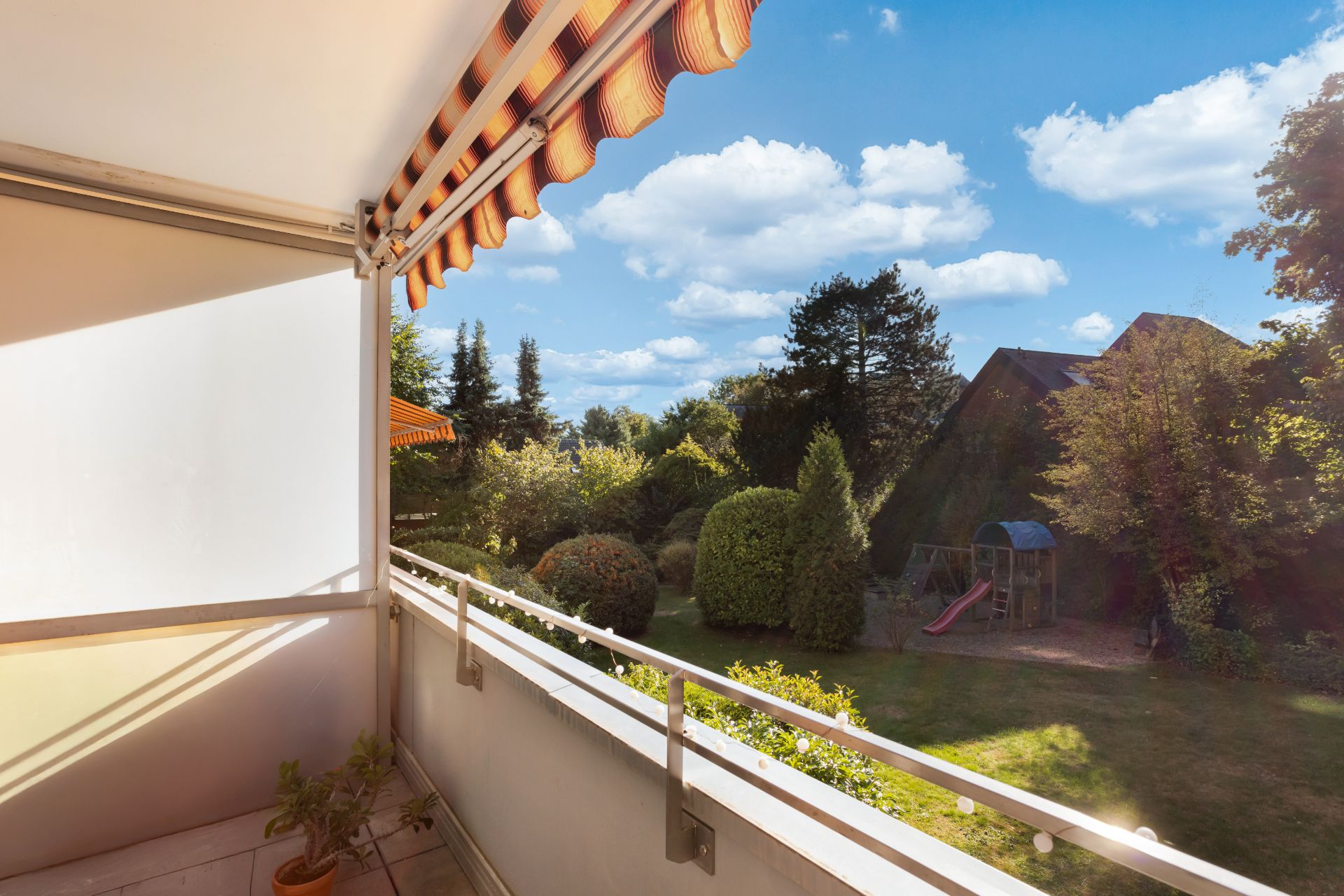 Balkon mit Markise, Blick auf Garten mit Rutsche und Bäumen bei sonnigem Wetter.