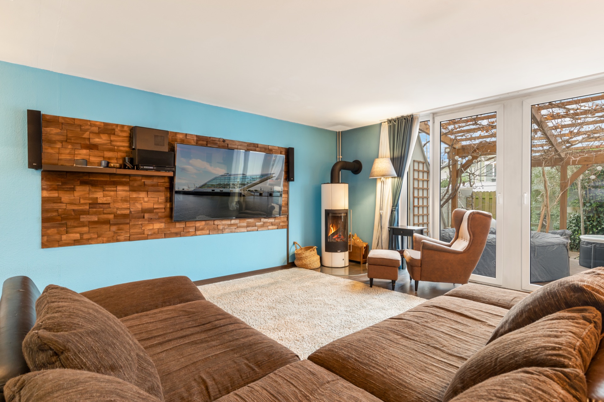 Wohnzimmer mit braunem Sofa, Kamin, Sessel, Fernseher und Blick auf den Garten.