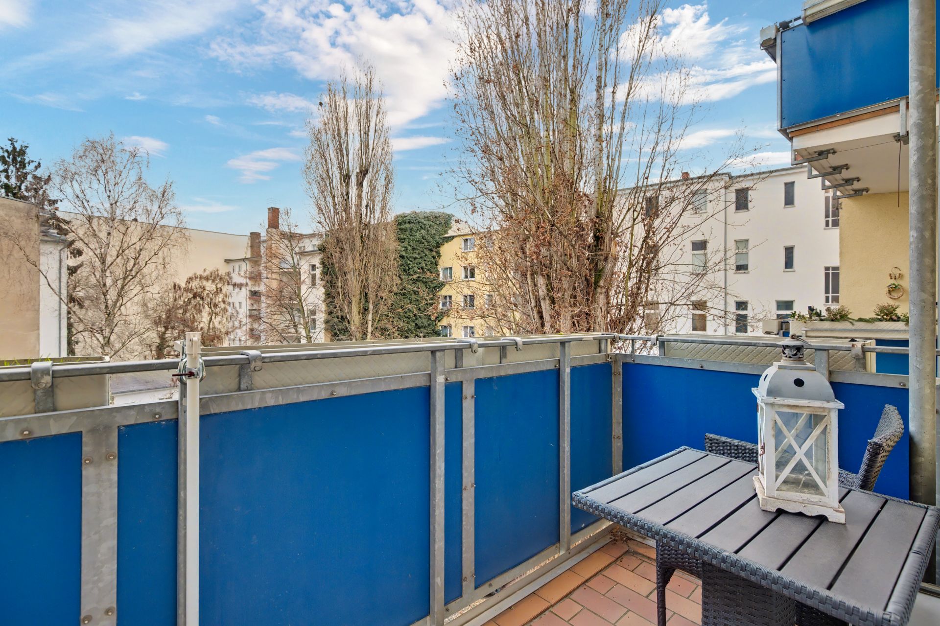 Balkon mit blauem Geländer, kleinem Tisch und Stuhl, Ausblick auf Bäume und Nachbargebäude.