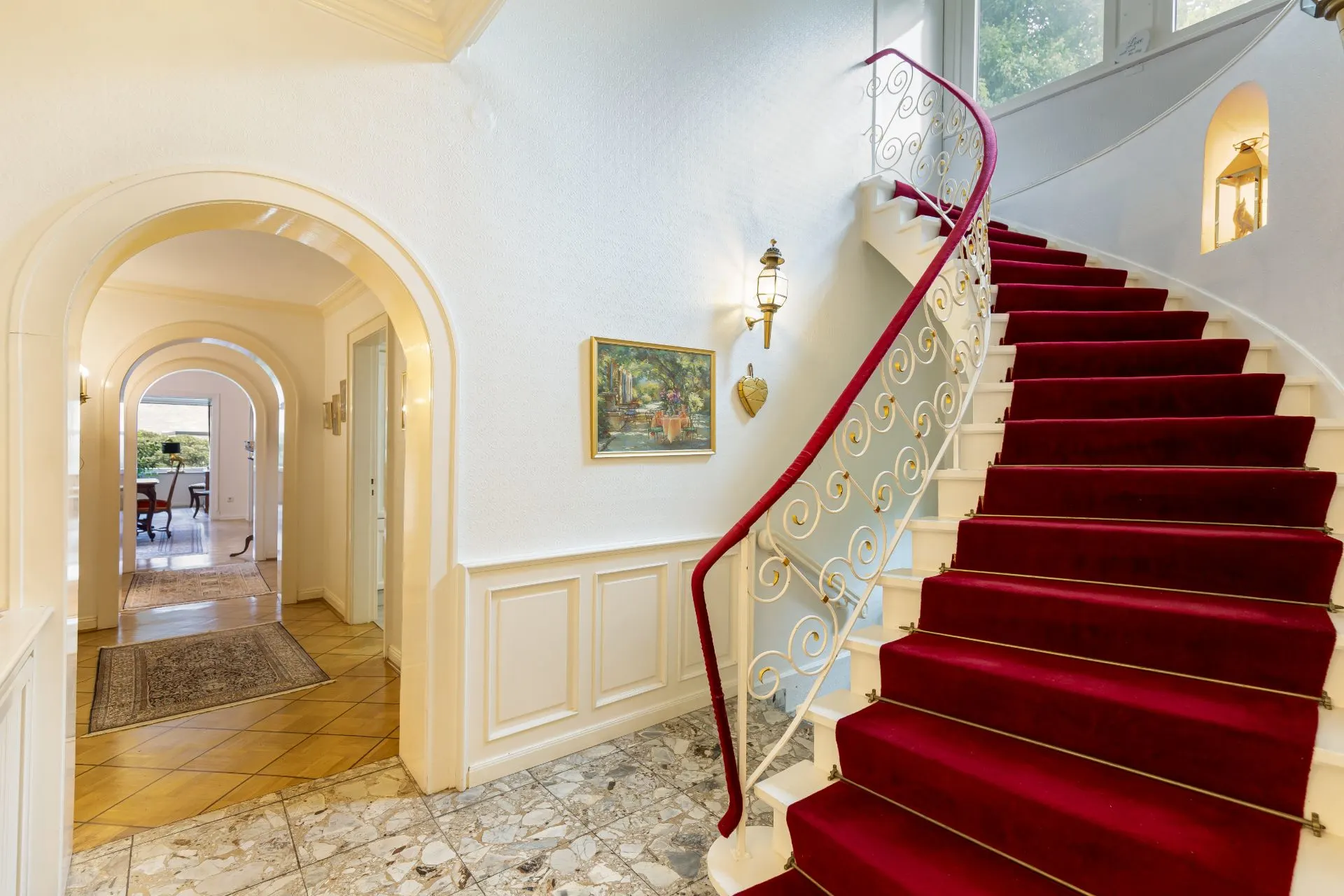 Treppenhaus mit rotem Teppich, geschwungene Treppe und heller Flur mit Wandbild.