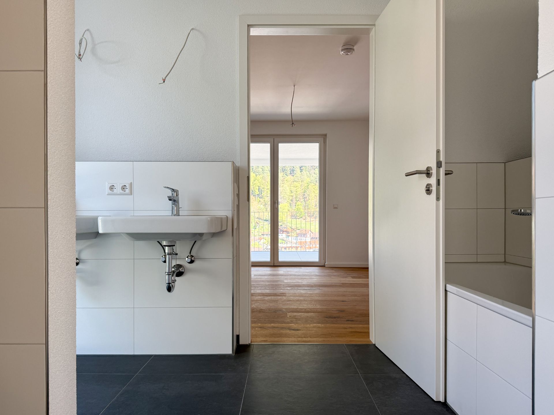 Helles Badezimmer mit Waschbecken, Badewanne und Blick in ein Zimmer mit Holzboden und Balkon.