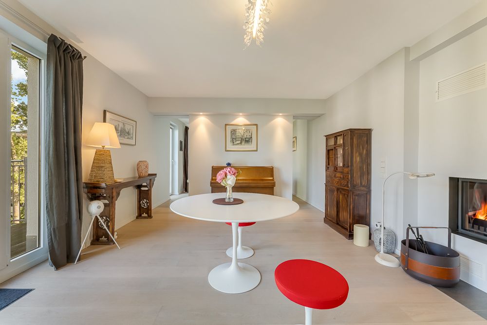 Helles Wohnzimmer mit rundem Tisch, zwei roten Hockern, Kamin und Balkonzugang.