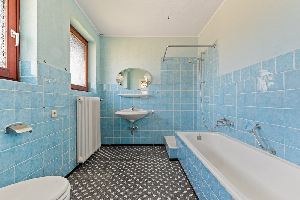 Badezimmer mit blauen Fliesen, Badewanne, Waschbecken, Spiegel und zwei Fenstern.