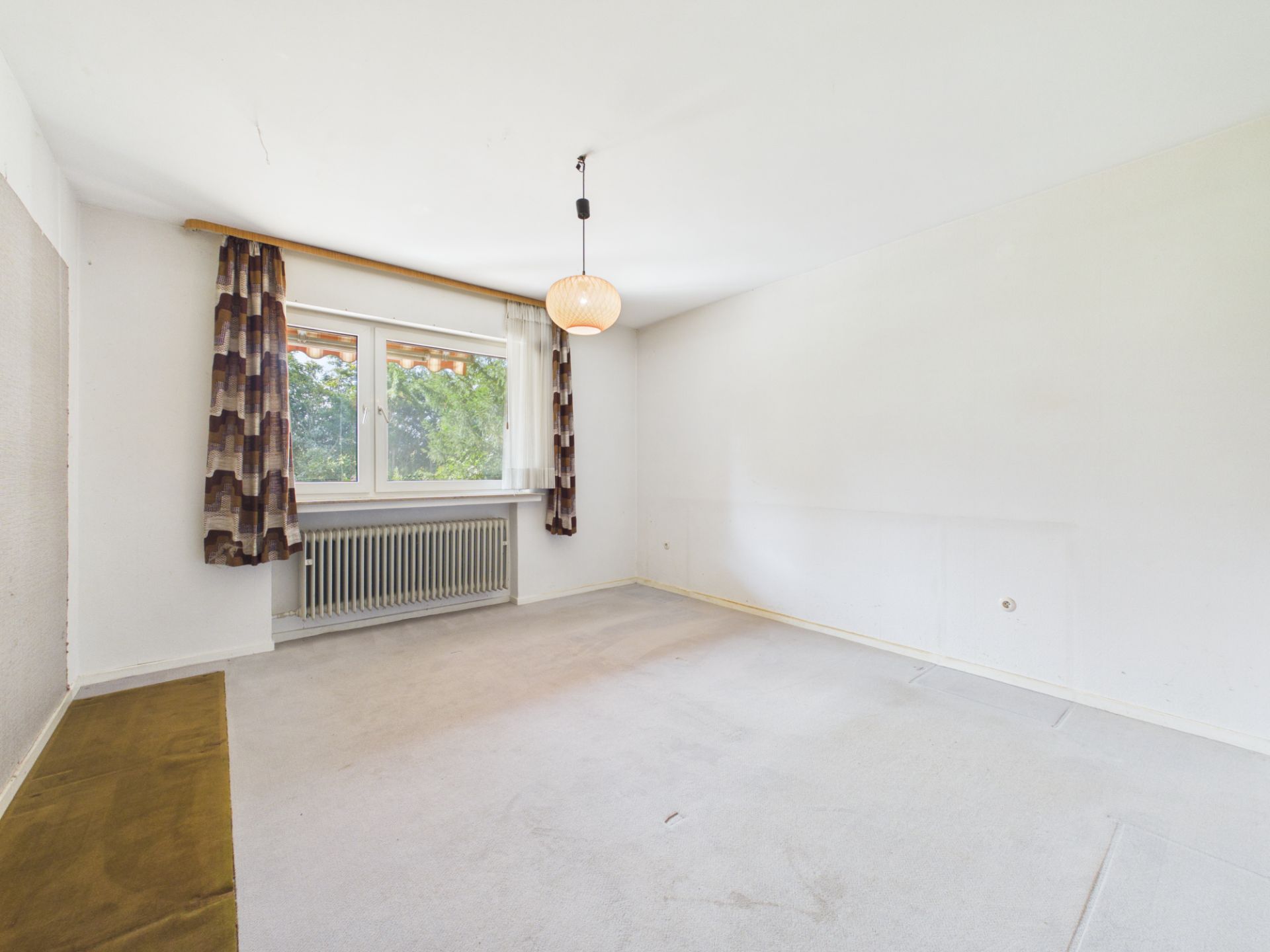 Helles, leeres Zimmer mit großem Fenster, Heizung und gemusterten Vorhängen.