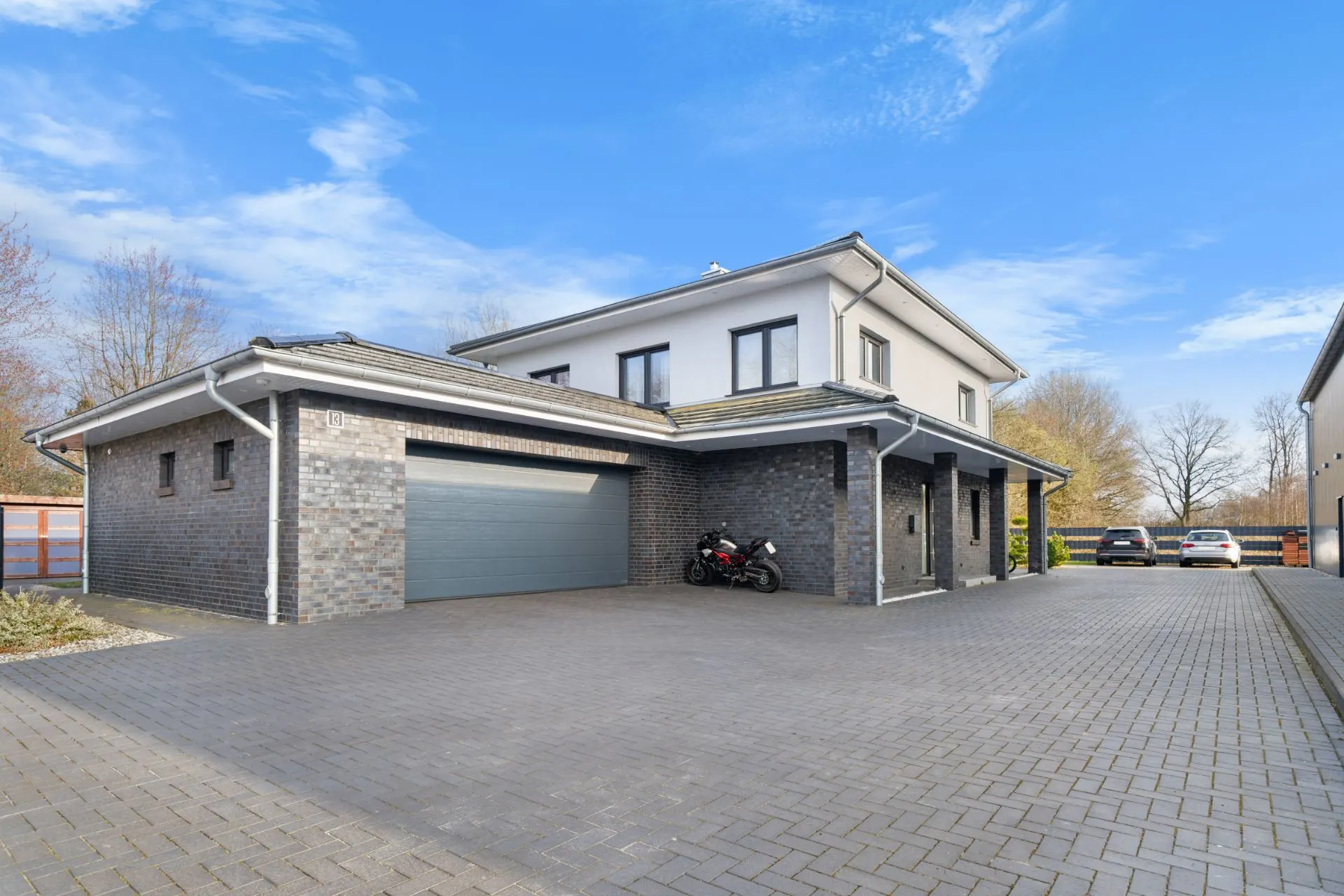 Modernes Wohnhaus mit Doppelgarage, Motorrad auf dem Hof und geparkten Autos im Hintergrund.