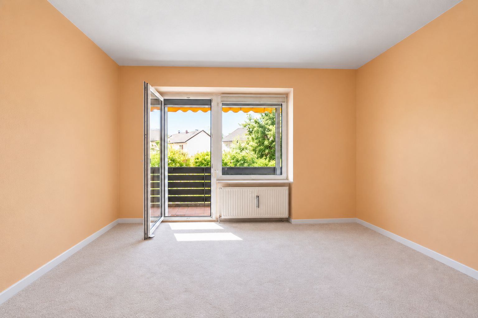 Leeres Zimmer mit beigen Wänden, Teppichboden und großer Tür zum Balkon mit Blick ins Grüne.
