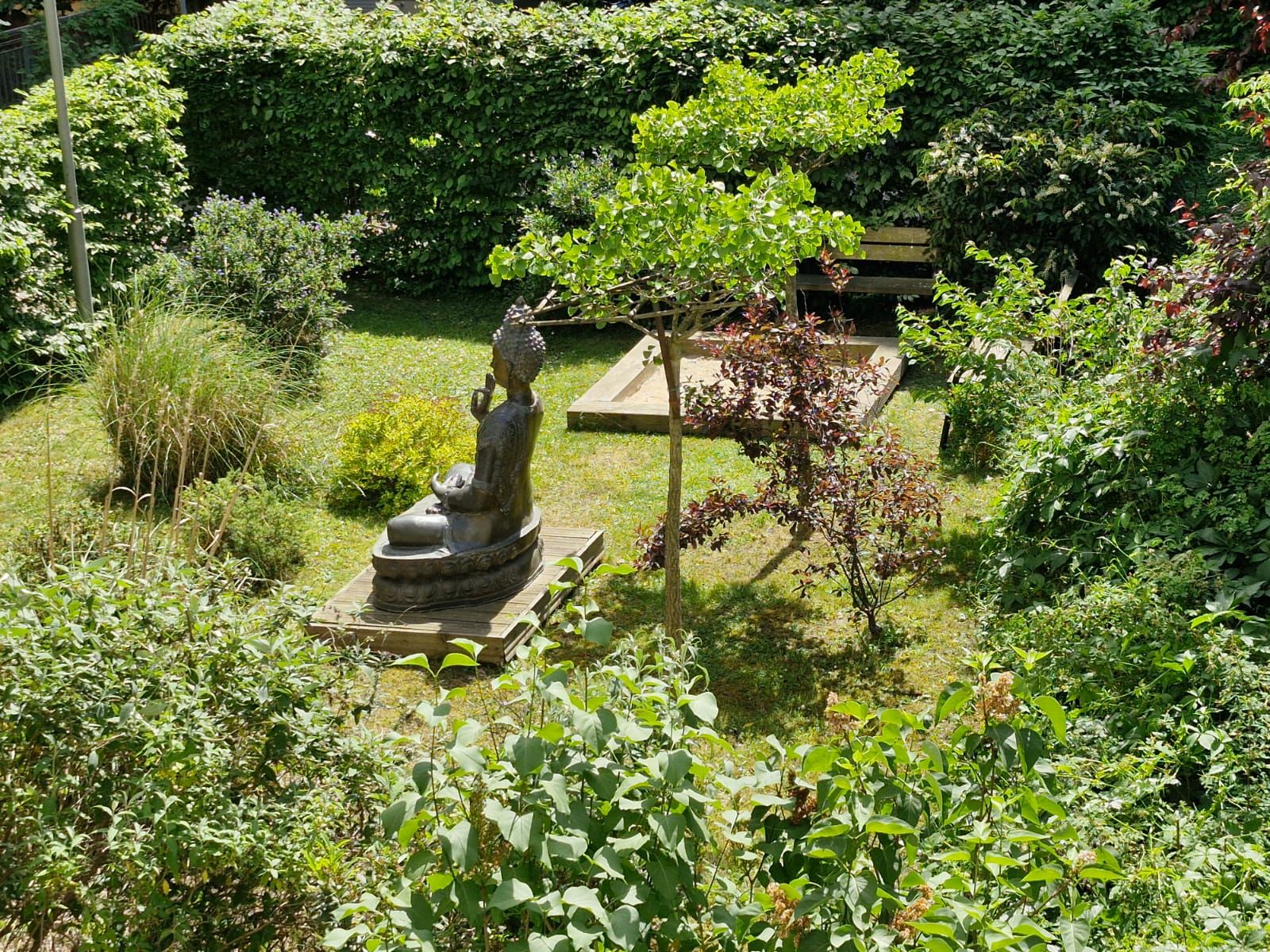 Garten mit einer Buddha-Statue, umgeben von Büschen, Bäumen und einer Holzbank im Hintergrund.