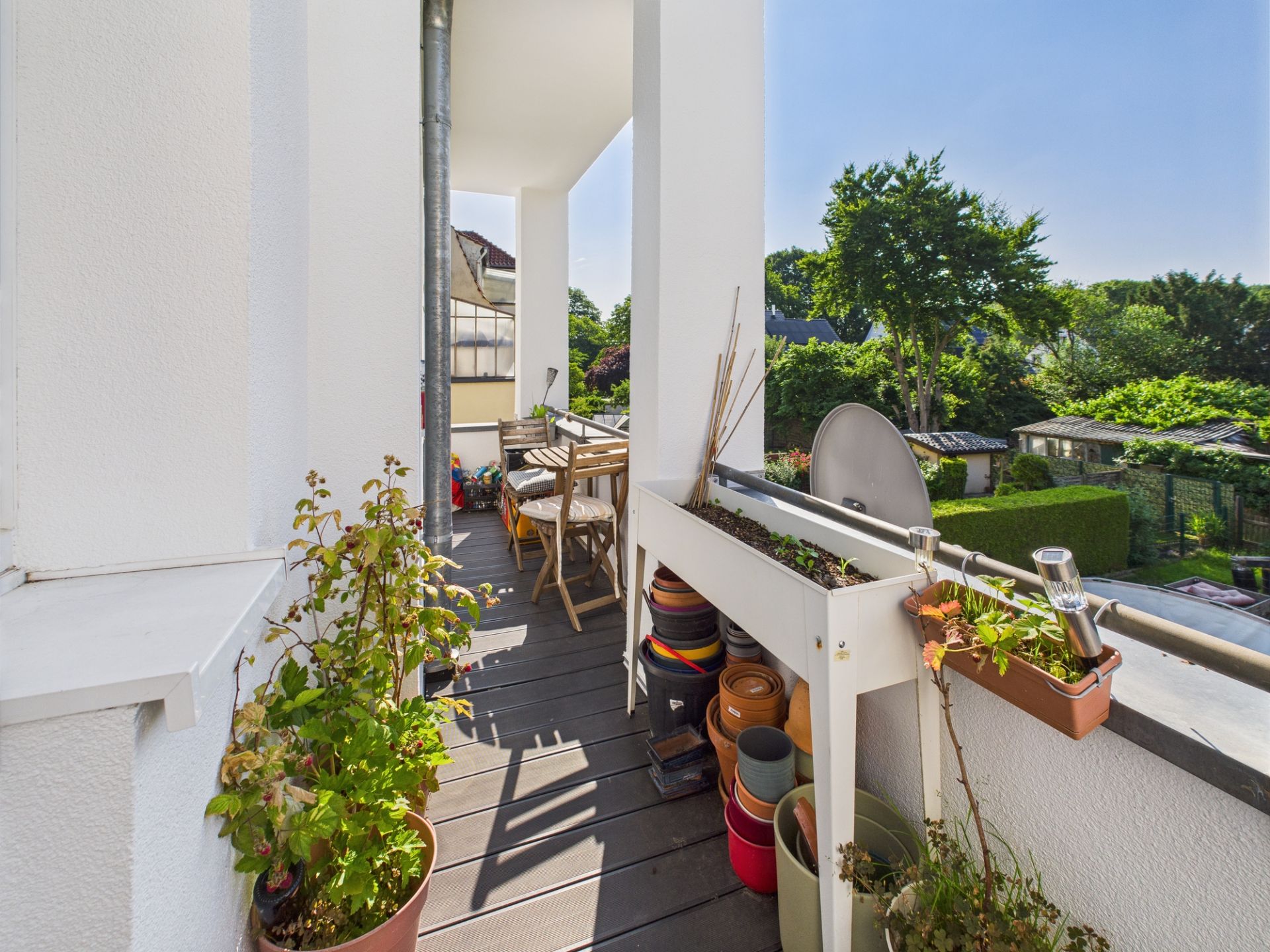 Balkon mit Pflanzen, Blumentöpfen und Holztisch mit Stühlen, Blick auf Garten und Bäume.