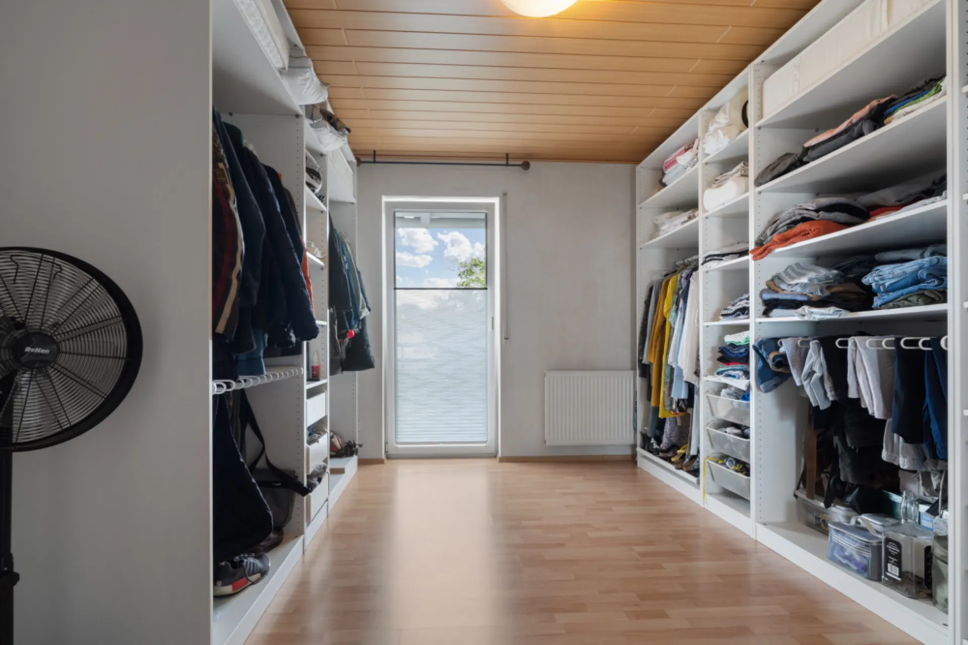 Großer begehbarer Kleiderschrank mit Regalen, Hängekleidung, gefaltetem Textil und einem Standventilator.