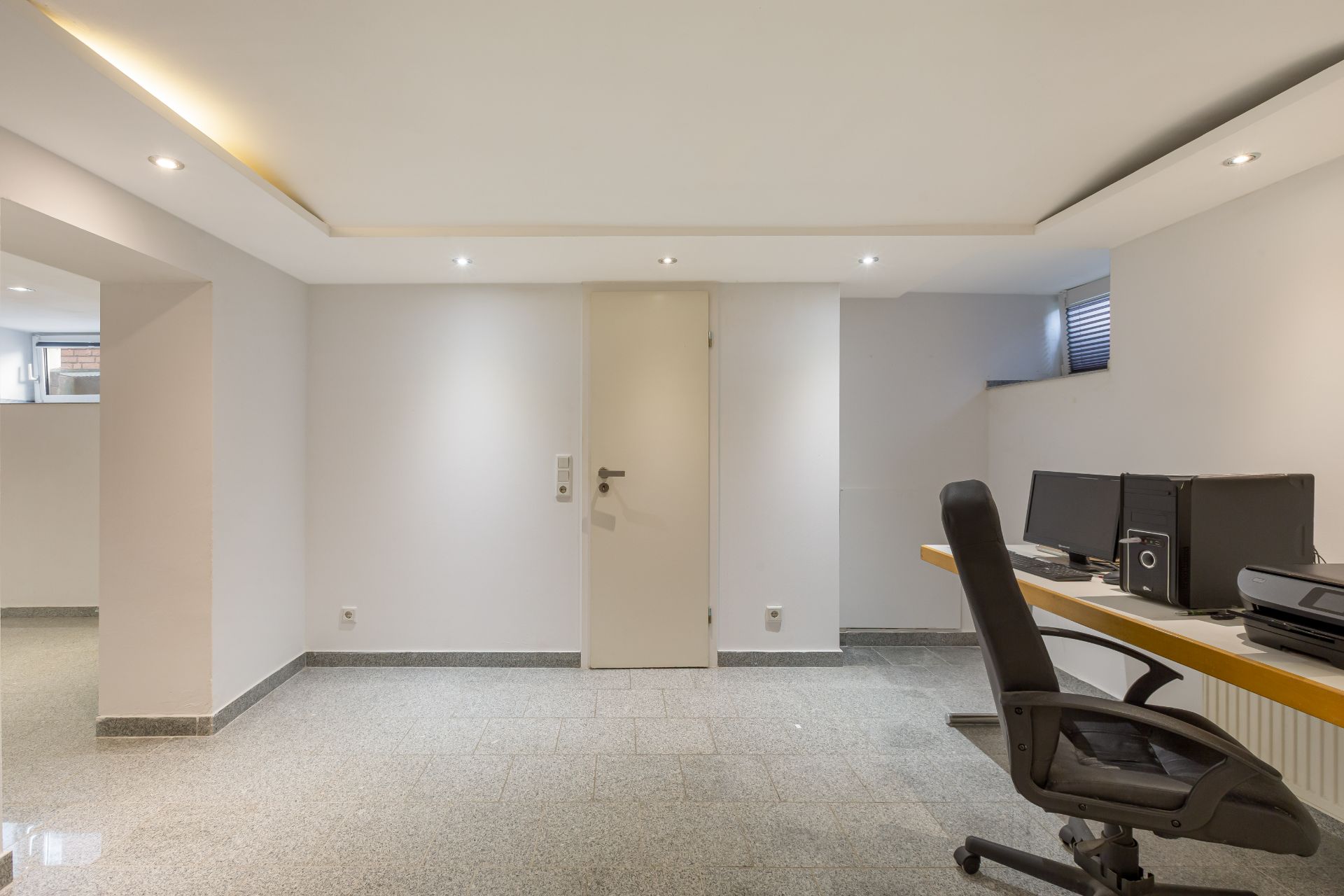Heller Kellerraum mit Schreibtisch, Bürostuhl und Computern, Tür in der Mitte, Fenster oben rechts.