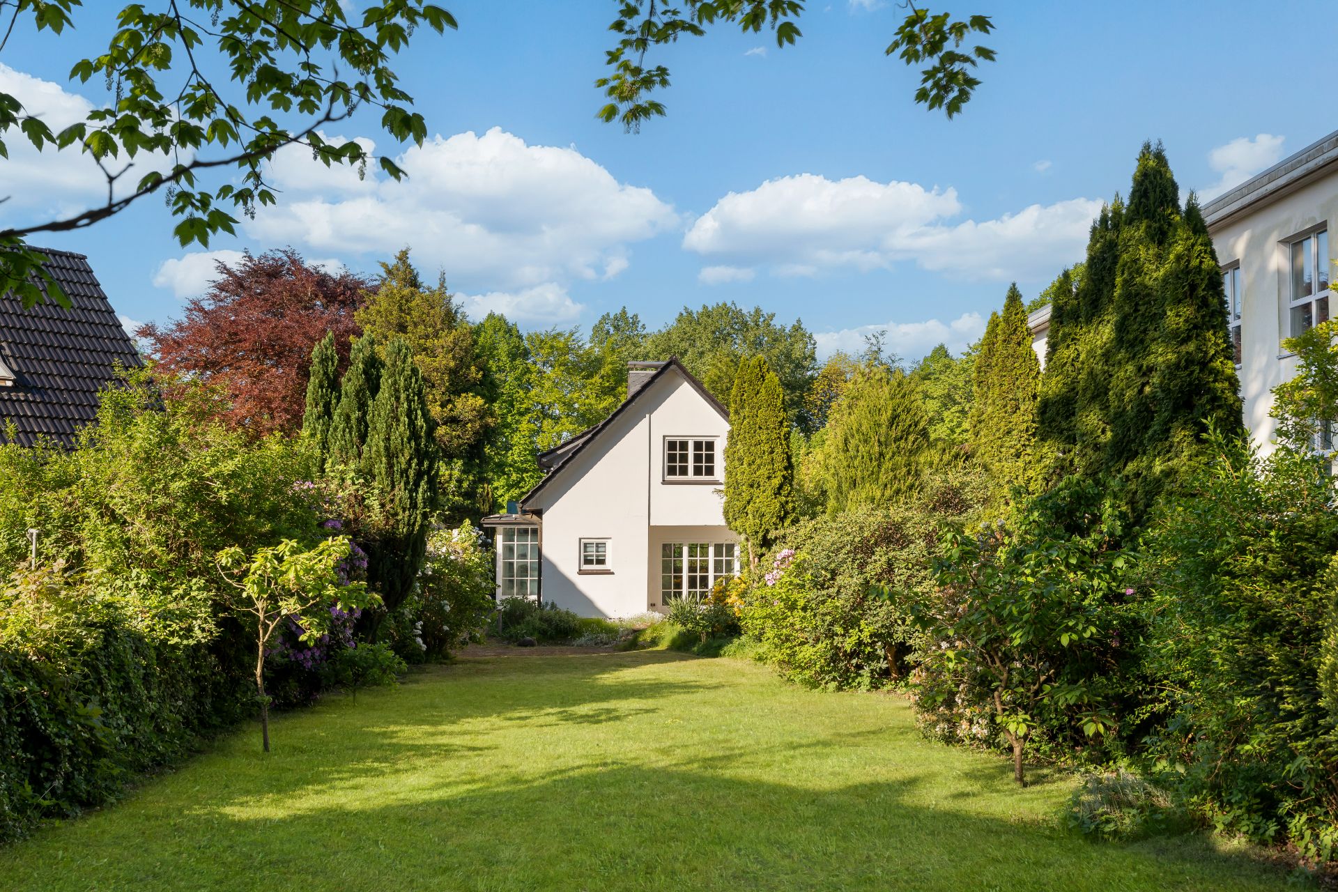 Weißes Haus mit Garten, umgeben von Bäumen und grüner Wiese, bei sonnigem Wetter.