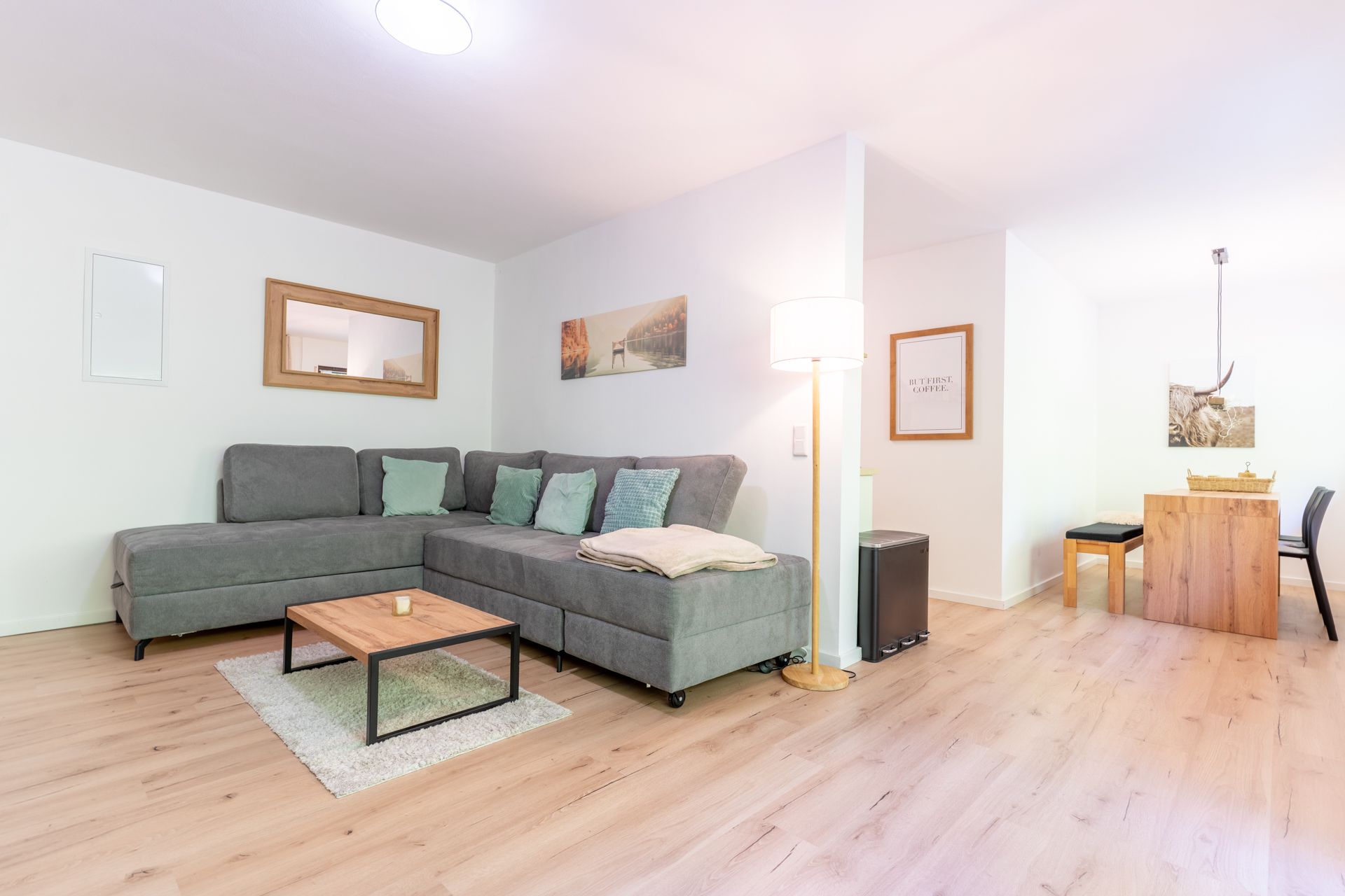 Helles Wohnzimmer mit grauem Ecksofa, Couchtisch, Stehlampe und Essbereich im Hintergrund.