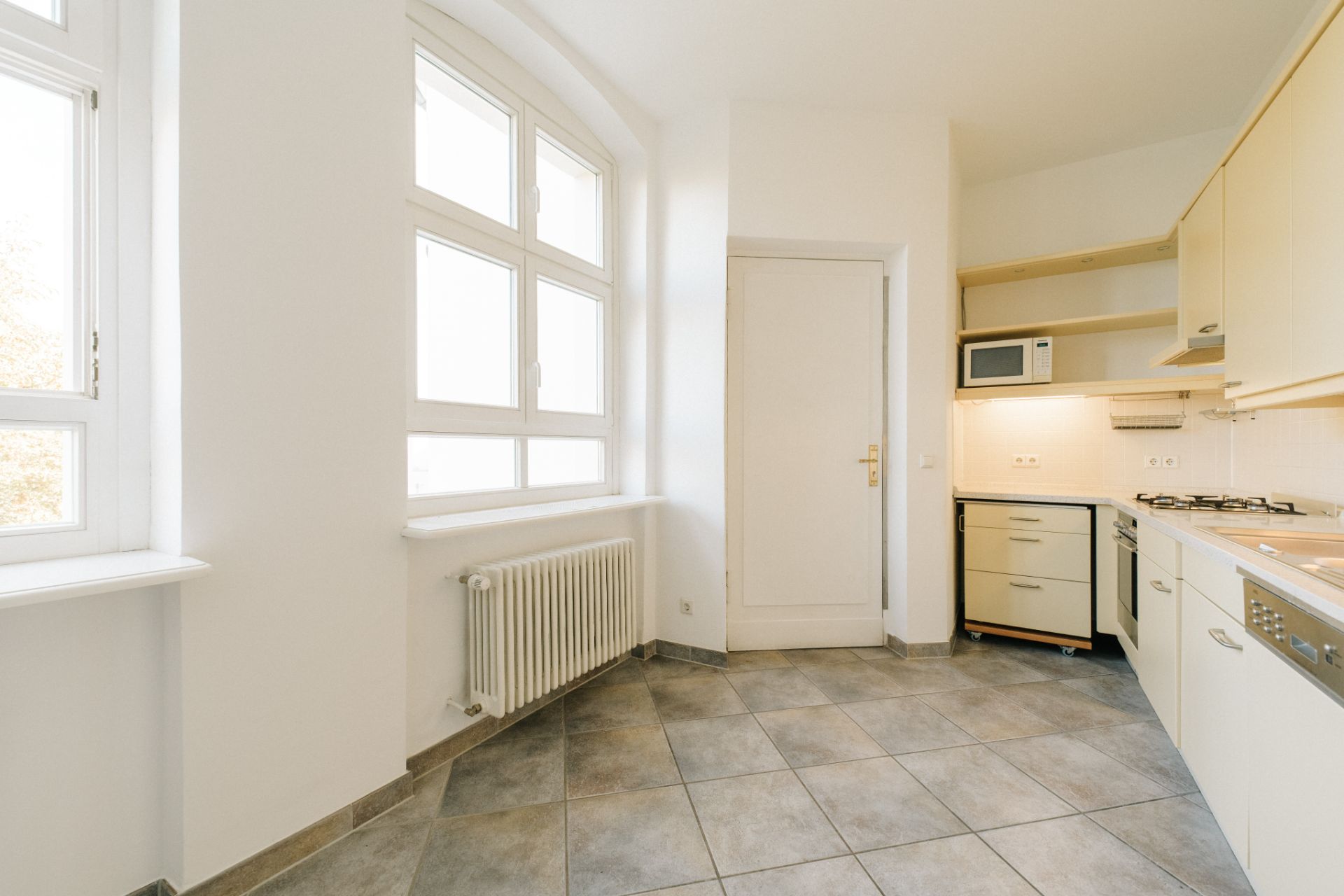 Helle Küche mit großen Fenstern, Fliesenboden, Heizkörper und Einbauküche mit Herd und Spülmaschine.