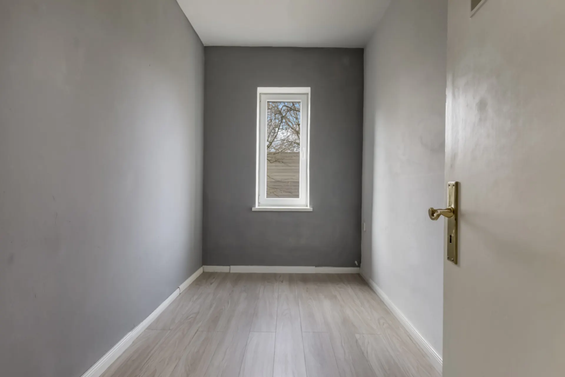 Kleiner leerer Raum mit grauen Wänden, Fenster und hellem Holzboden.