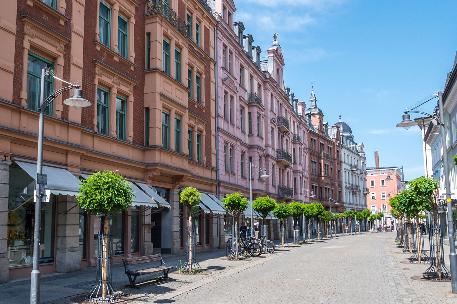 Breite Straße mit alten Wohnhäusern, Bäumen, Bänken und Fahrrädern bei Sonnenschein.