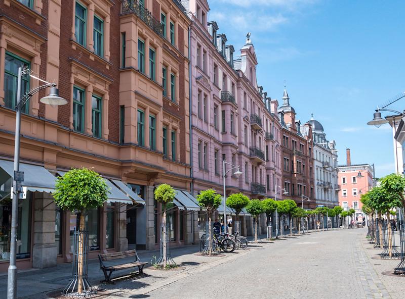 Breite Straße mit alten Wohnhäusern, Bäumen, Bänken und Fahrrädern bei Sonnenschein.